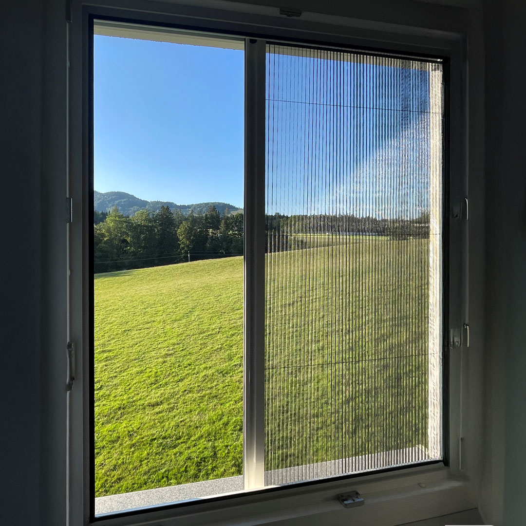 halb geöffnetes Insektenschutzplissee an einem Fenster mit Blick auf Wiese.