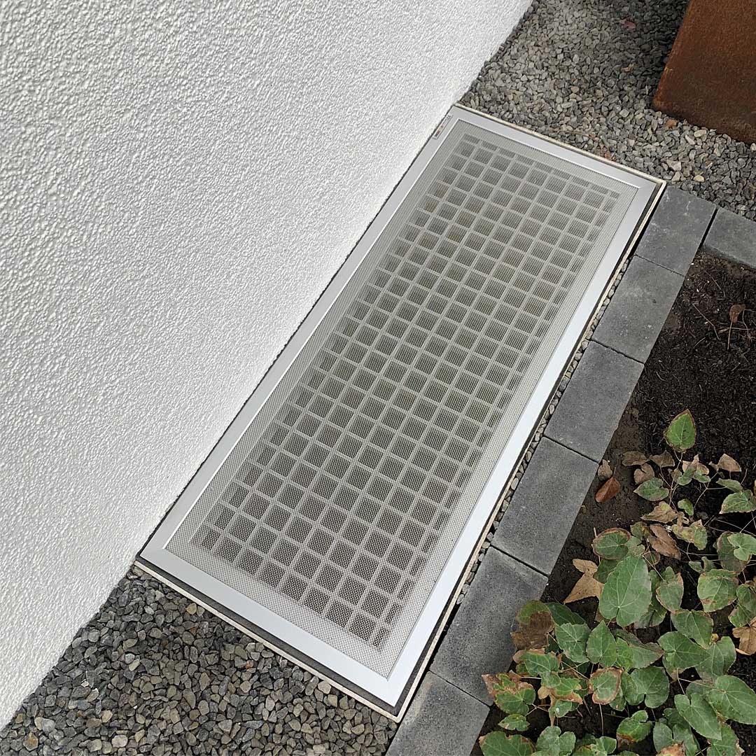 Lichtschachtabdeckung liegt fest neben Kies und Hauswand.