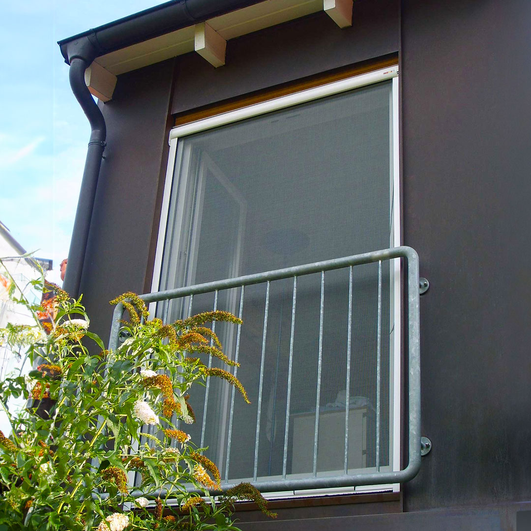 Fenster mit Insektenschutzrollo hinter Balkongeländer und Pflanzen