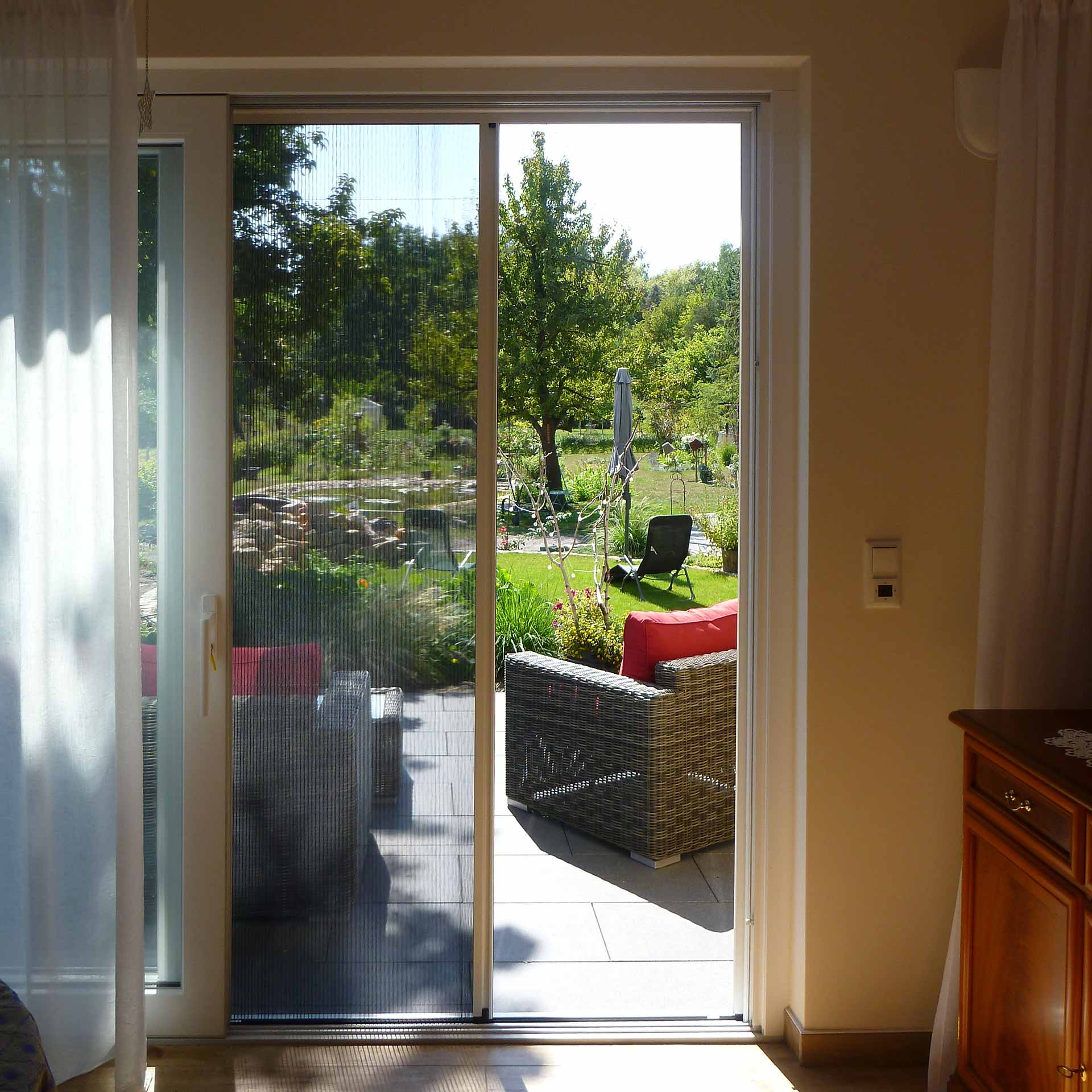 Fliegengittertür trennt Wohnzimmer von sonnigem Garten mit Möbeln.