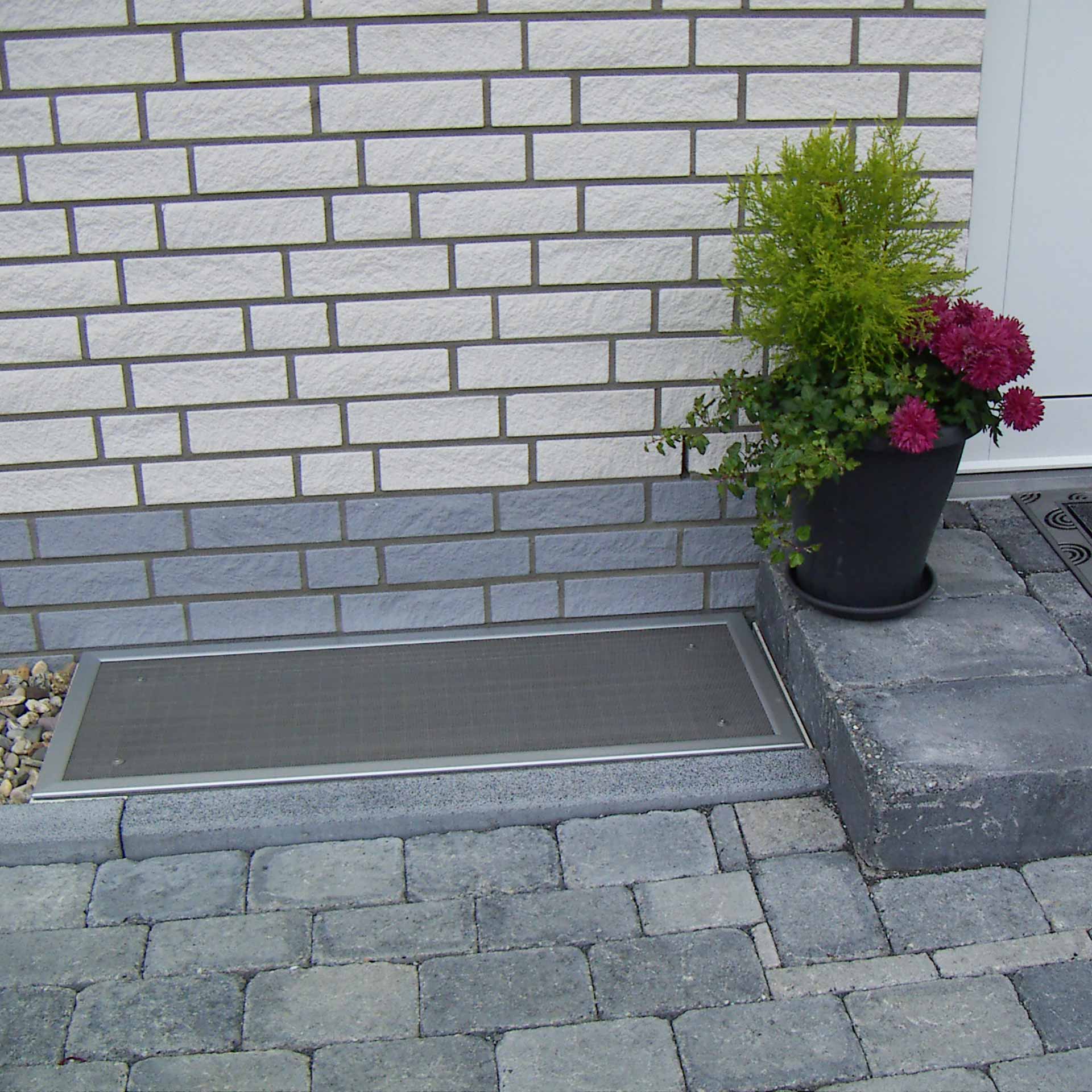 Lichtschachtabdeckung auf Maß für Ihren Kellerschacht | Cleanline Lichtschachtabdeckung schützt Kellerfenster, vor Wand mit Pflanzentopf.