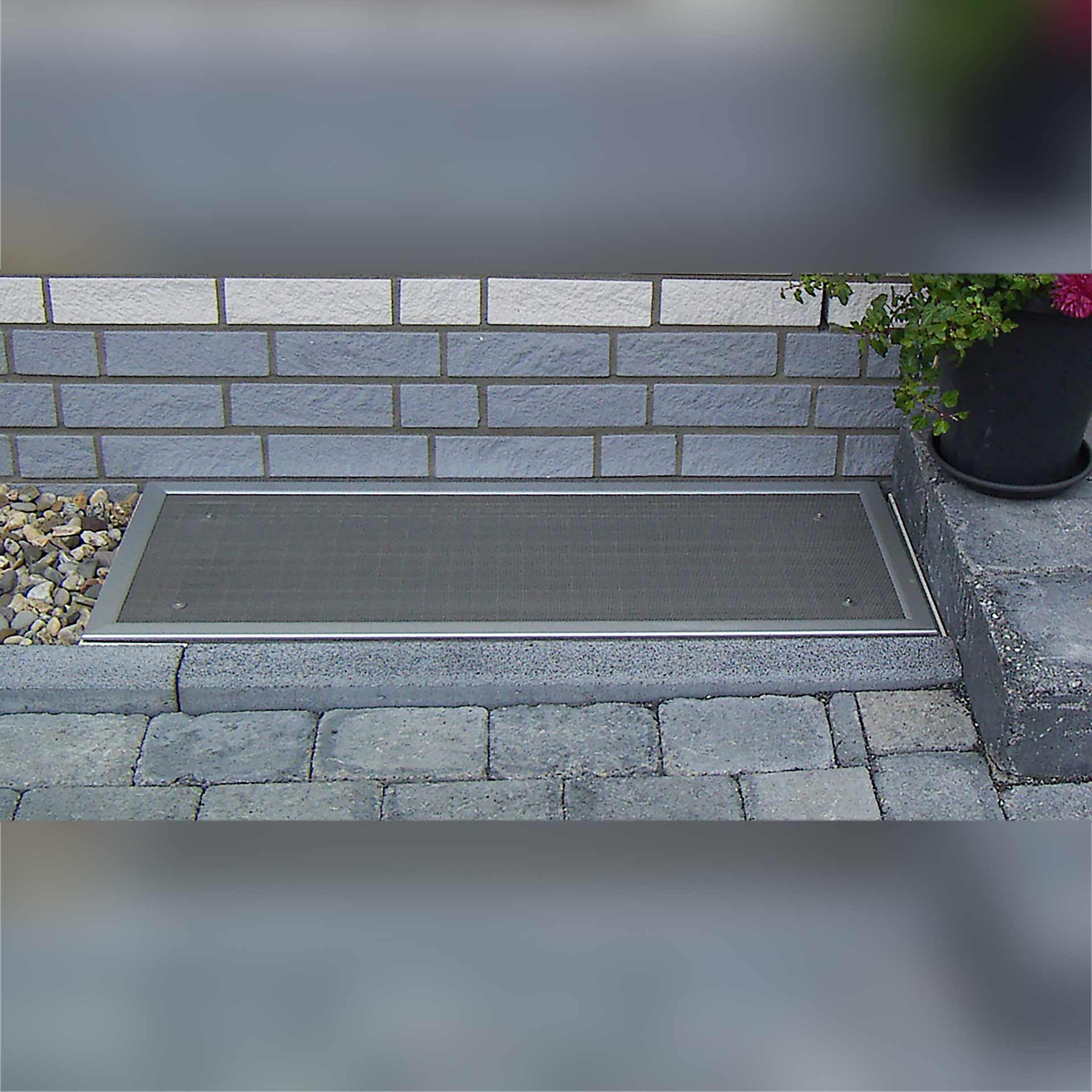 Lüftungsschachtabdeckung, vor gemauerter Hauswand auf Steinboden.