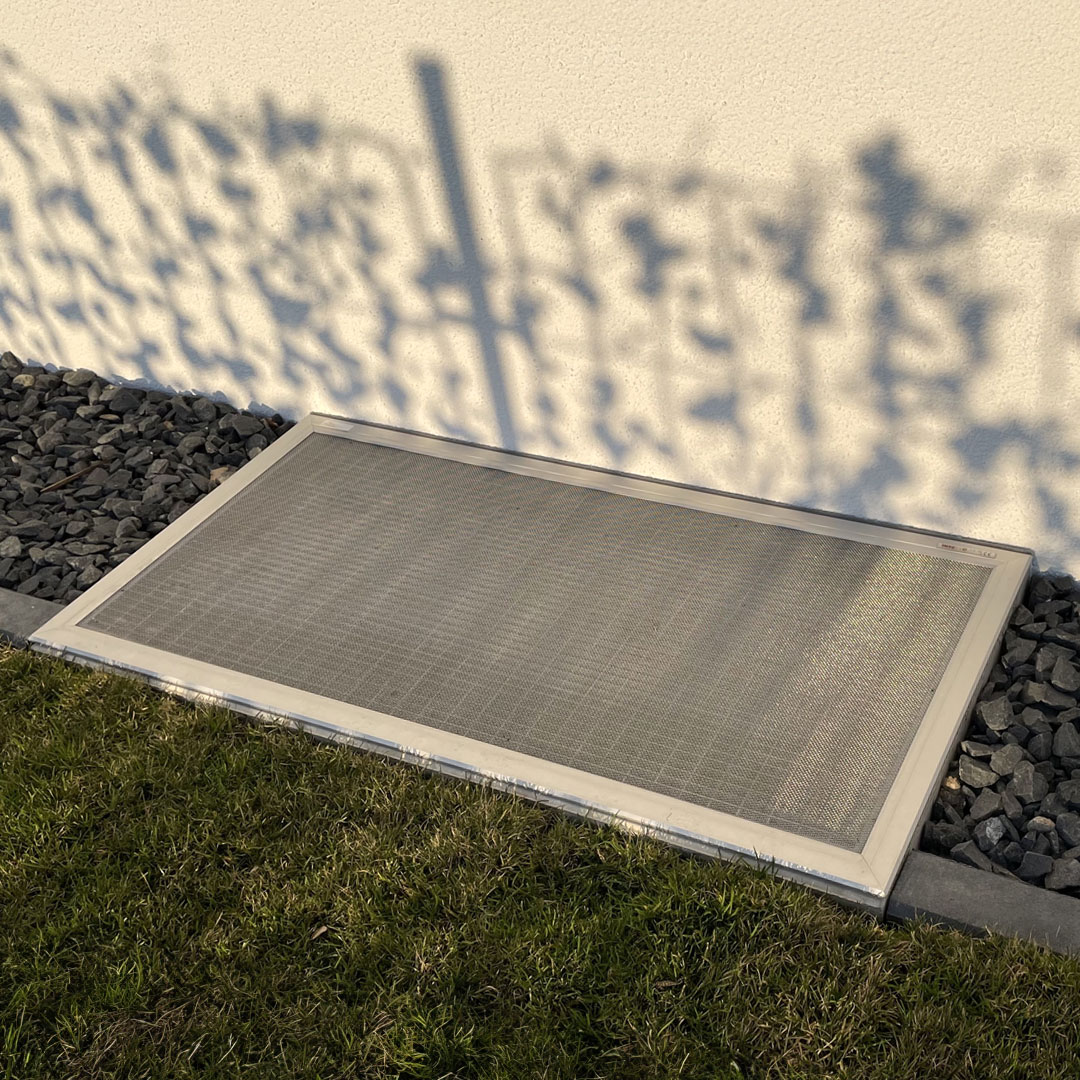 Insektenschutzgitter bedeckt Lichtschacht, neben Steinbeet und Rasenfläche.