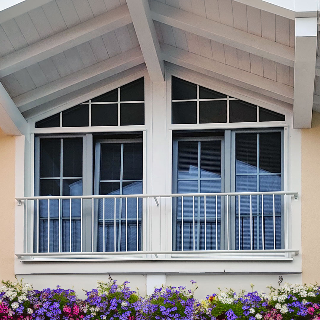 Fenster spiegelt Himmel, umrahmt von weißem Dach, Blumen unten.