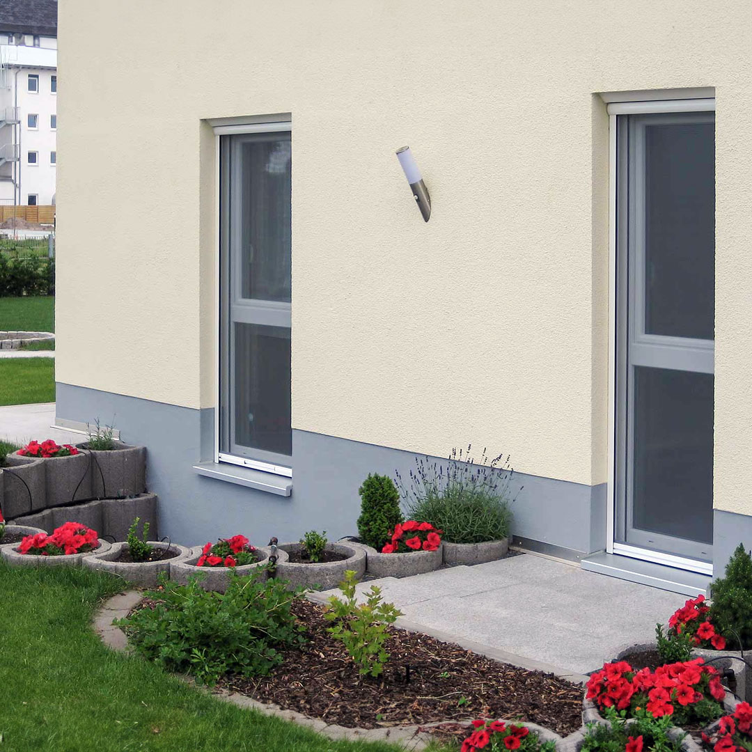 Fenster, ausgestattet mit Fliegengitter, in Blumenbeet umgebenem Haus.