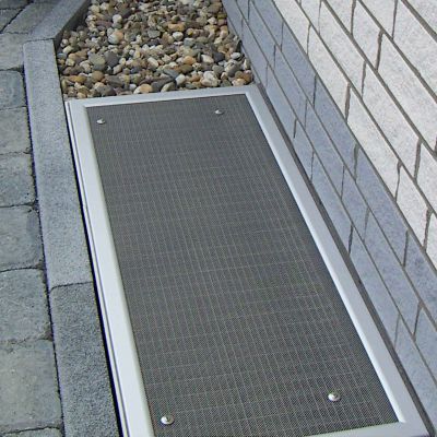 Lichtschachtabdeckung , umgeben von Kies, Pflastersteinen und Ziegelwand