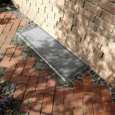 Lichtschachtabdeckung liegt am Boden neben einer Ziegelmauer.